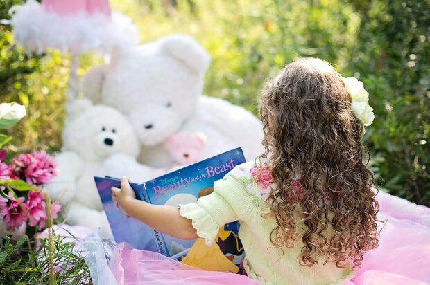 Prikaz devojčice koja čita bajku i sedi na livadi sa plišanim igračkama ispred nje